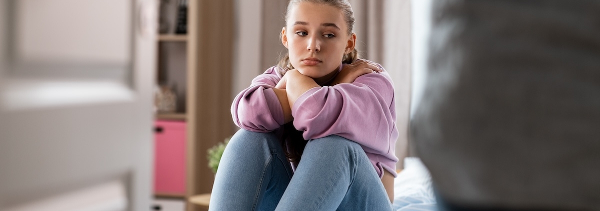 Sad teen girl sits on bed while parent enters room