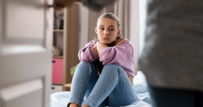 Sad teen girl sits on bed while parent enters room