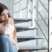 Teen girl sits on steps inside metal railing, backpack at feet, looking burdened